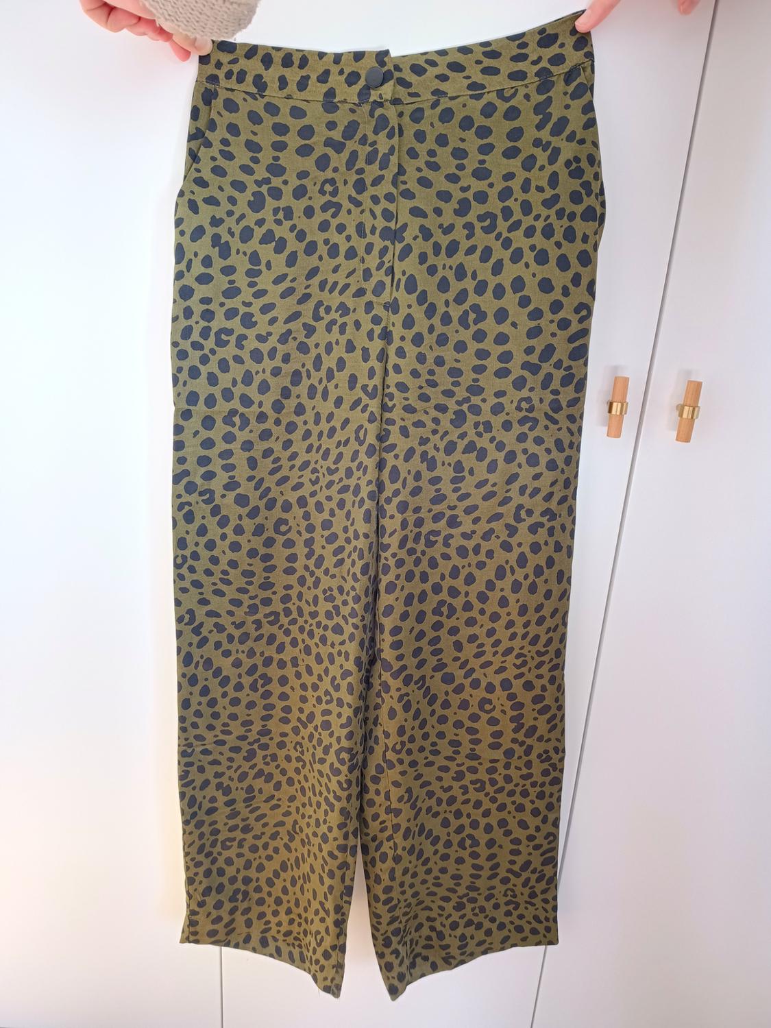 Green fabric trousers with a black spot pattern, held by two hands against a white wardrobe background.