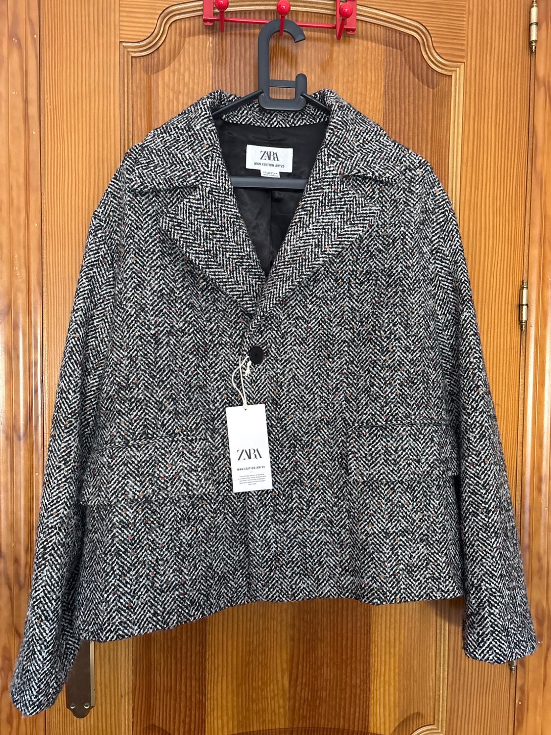 Grey jacket with a herringbone pattern, hanging on a wooden door. Visible label with the text "ZARA WOMAN EDITION 1975".