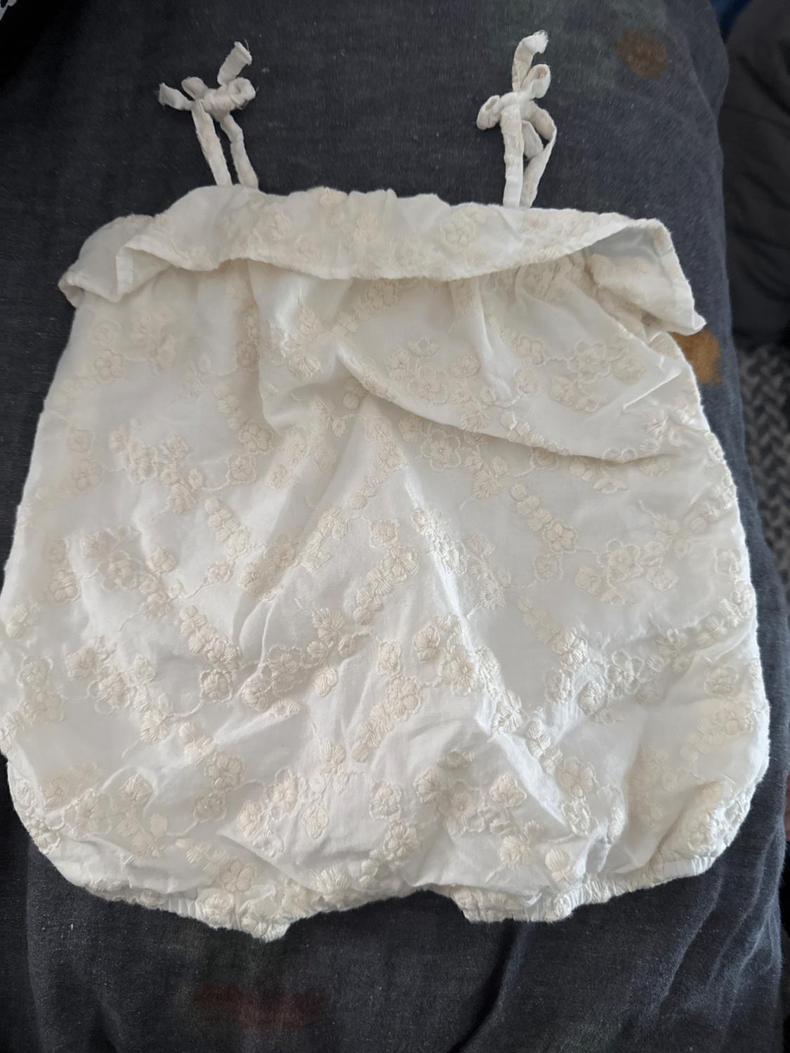 White children's garment with bow straps and floral embroidery on a dark grey background.