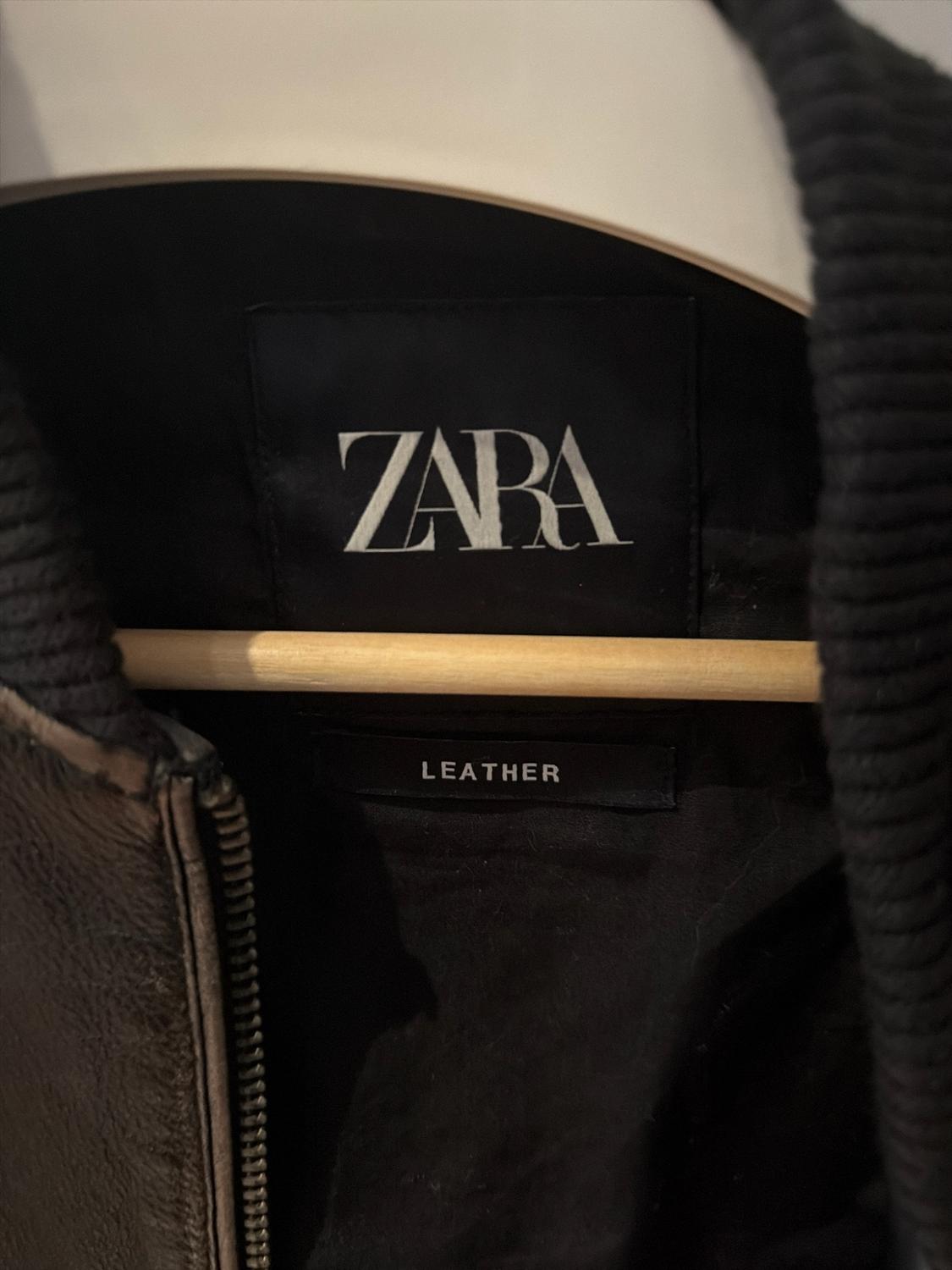 Close-up of the inside of a dark jacket on a wooden hanger. The labels with the text ZARA and LEATHER are visible.