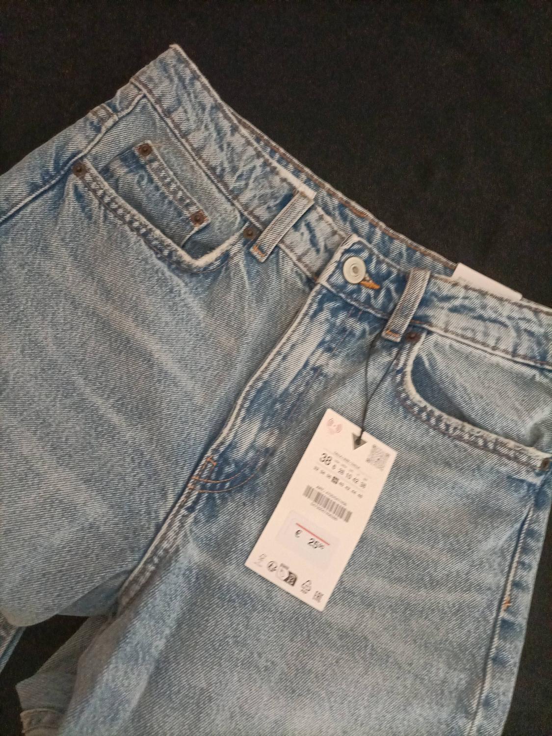 Close-up of light blue jeans on a dark background. It shows the waistband, pockets, and a label with the text 38 and €25.95.
