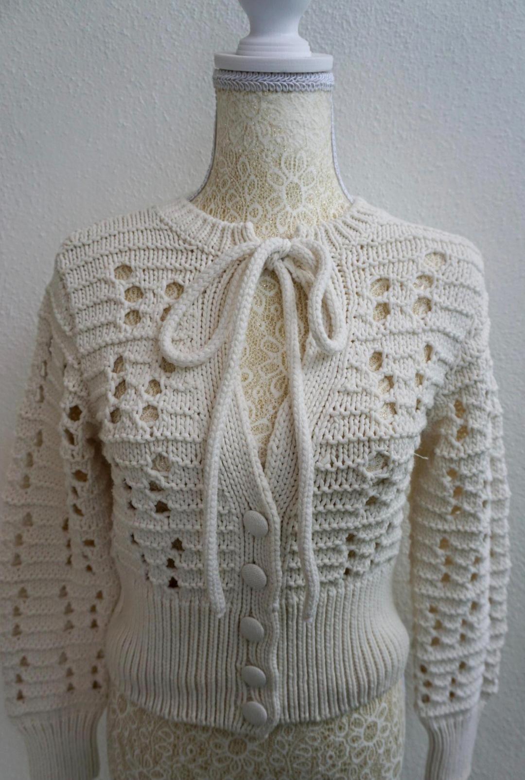 Mannequin wearing a white knitted cardigan with openwork details, front buttons and a bow at the neck, against a light background.