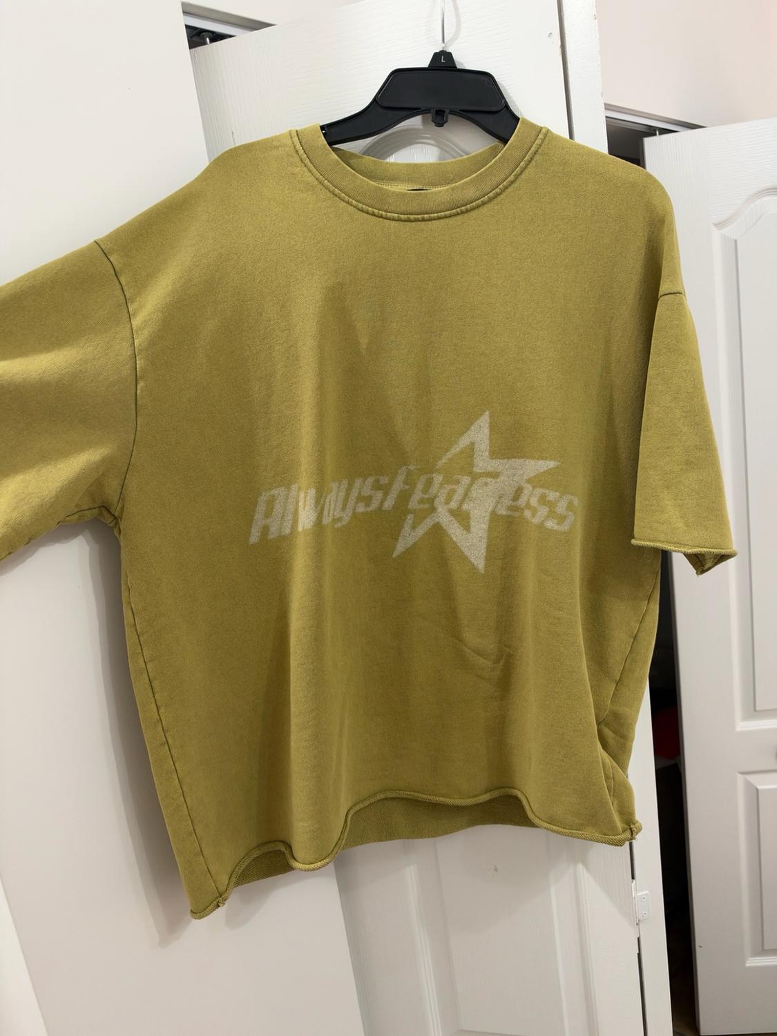 Olive green crewneck T-shirt, hanging on a black hanger. It has a “Always fearless” graphic with a star in the center.