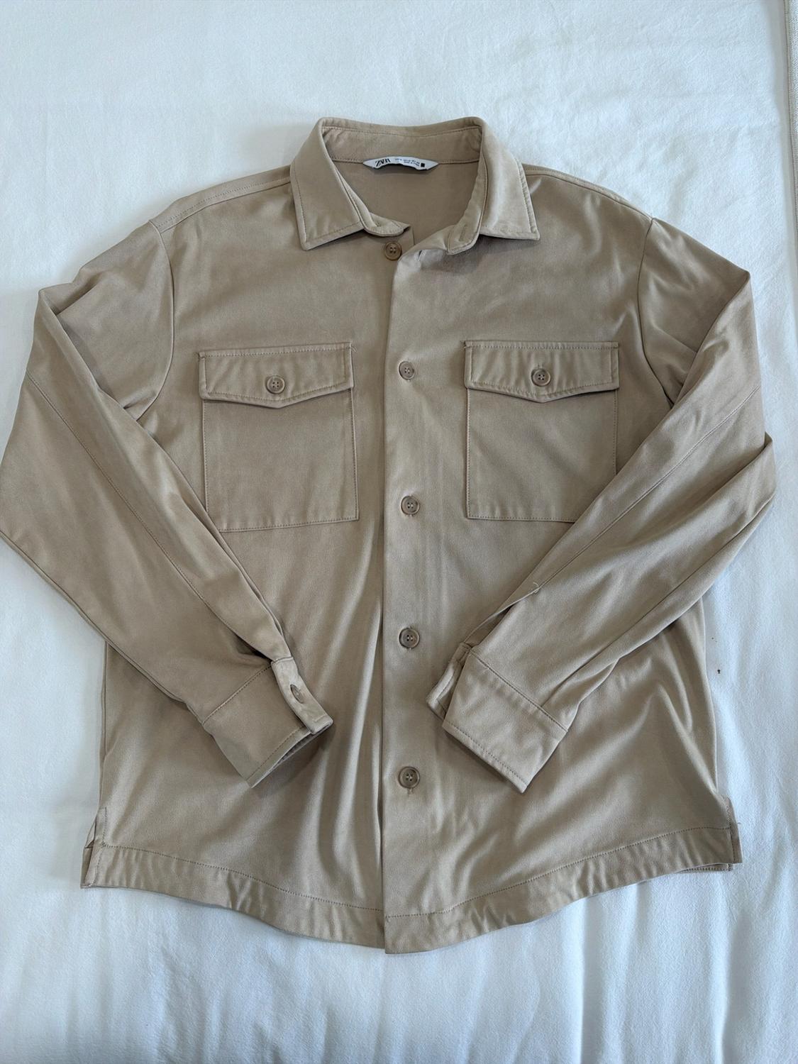 Beige long-sleeved button-down shirt with two front pockets, laid flat on a white fabric surface. ZARA label visible.