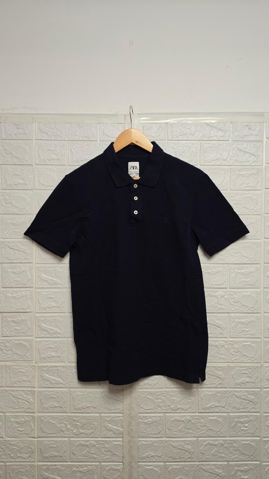 Black polo shirt hanging on a wooden coat hanger on a white tiled wall. Label visible with the text "ZARA".