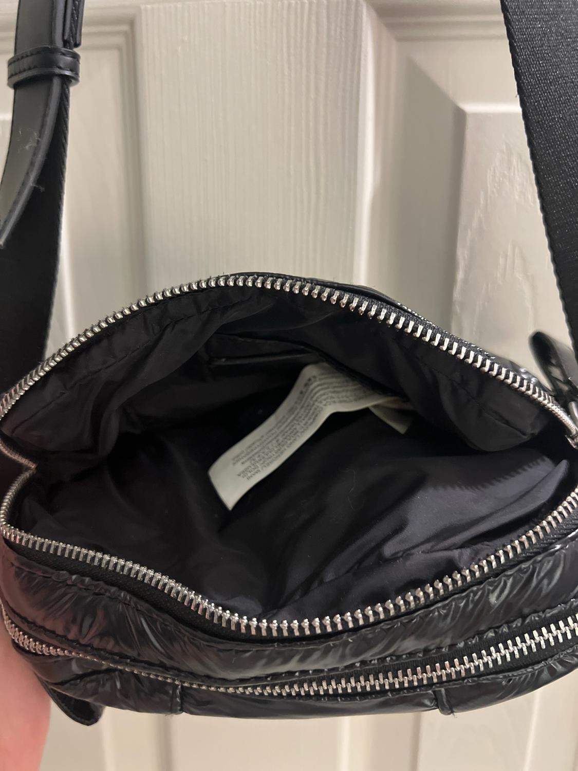 Open quilted black bag with a silver zipper, showing its dark interior and white label. Black strap and white door background.