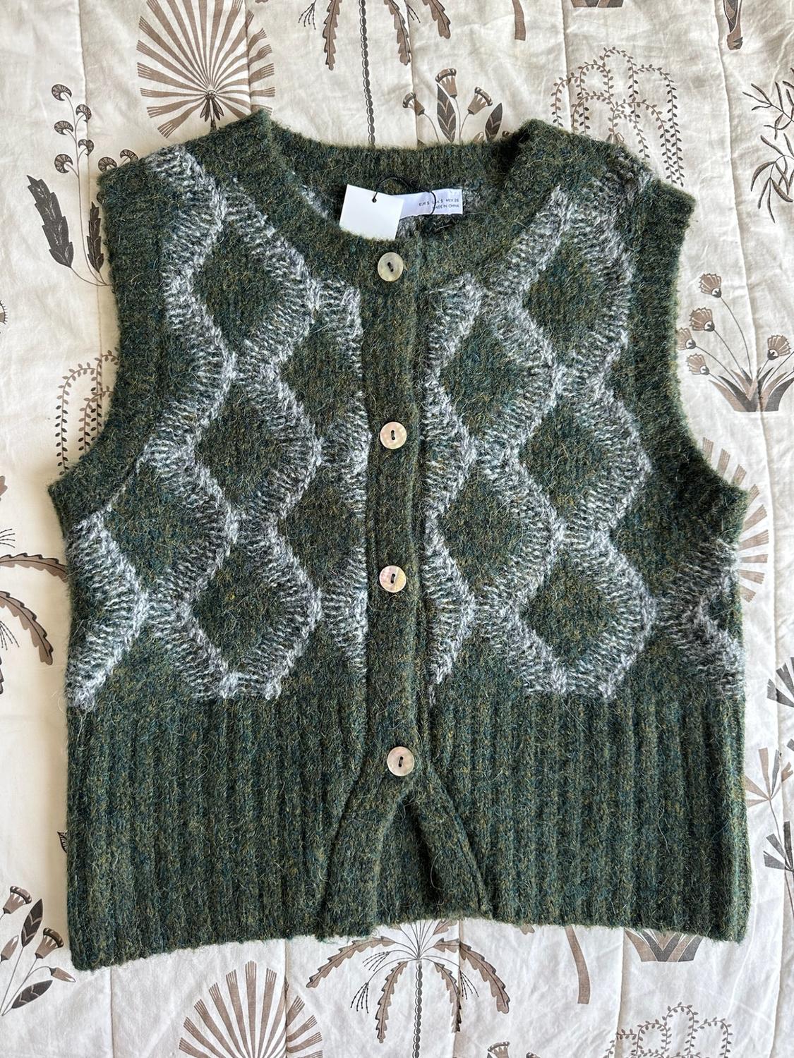 Green knitted waistcoat with white diamonds and front buttons, placed on a light-coloured fabric with a botanical print.