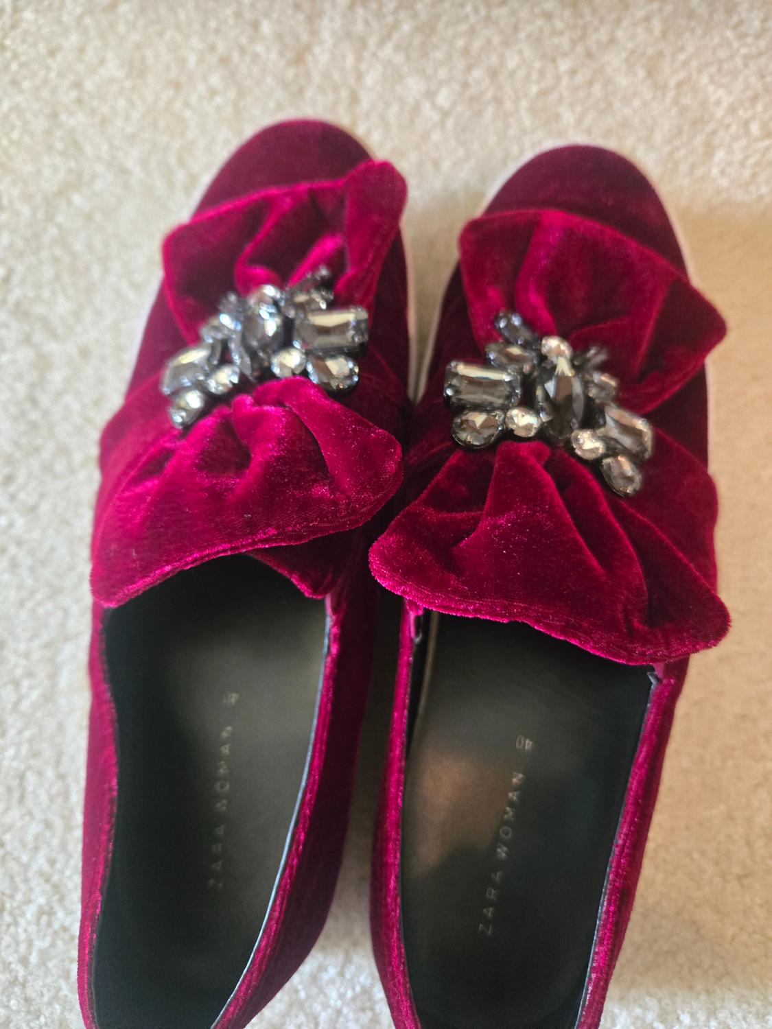 Red velvet shoes with bow and rhinestones on the upper. Black interior with "ZARA WOMAN" text. Beige carpet background.