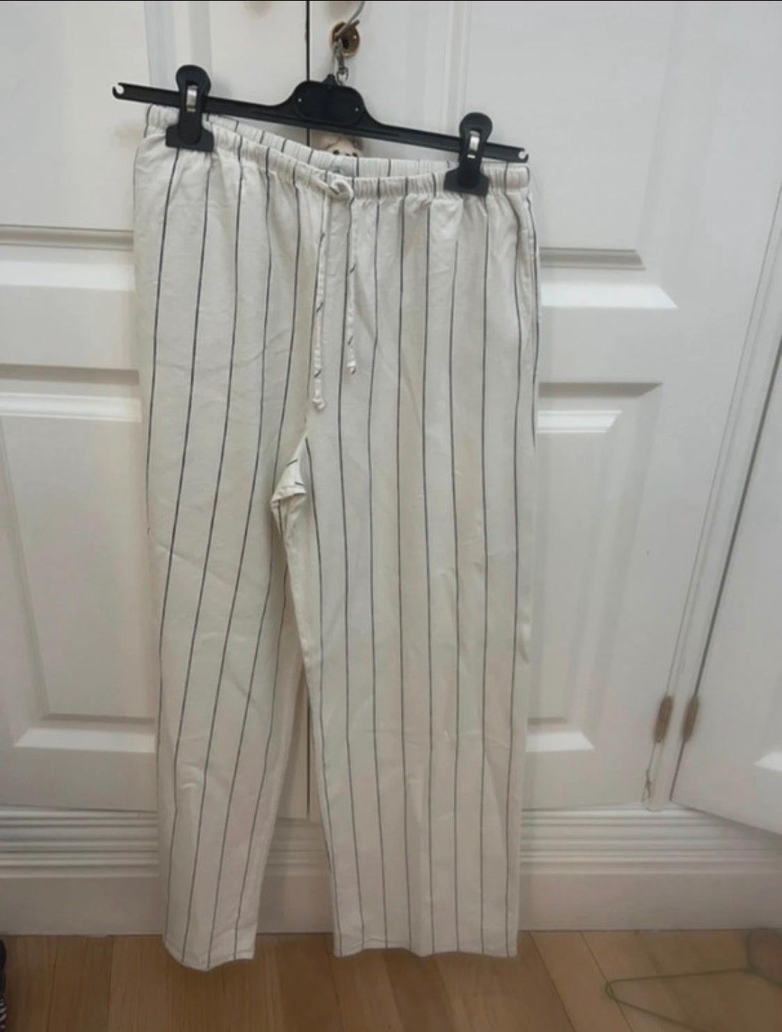 Light-coloured trousers with dark vertical stripes and a drawstring, hanging on a hanger in front of a white door. Casual style and relaxed vibe.