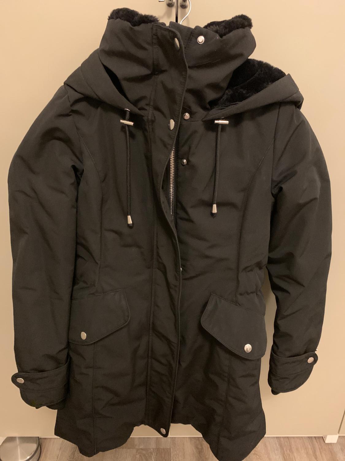 Black coat with a hood and plush lining, hanging on a door. It has flap pockets and a zip fastening.