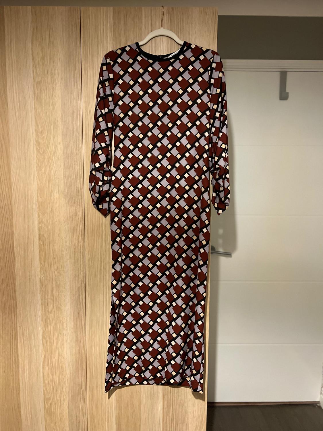 Long dress with a geometric print in brown, white, and black tones, hanging on a hanger against a light wooden background.