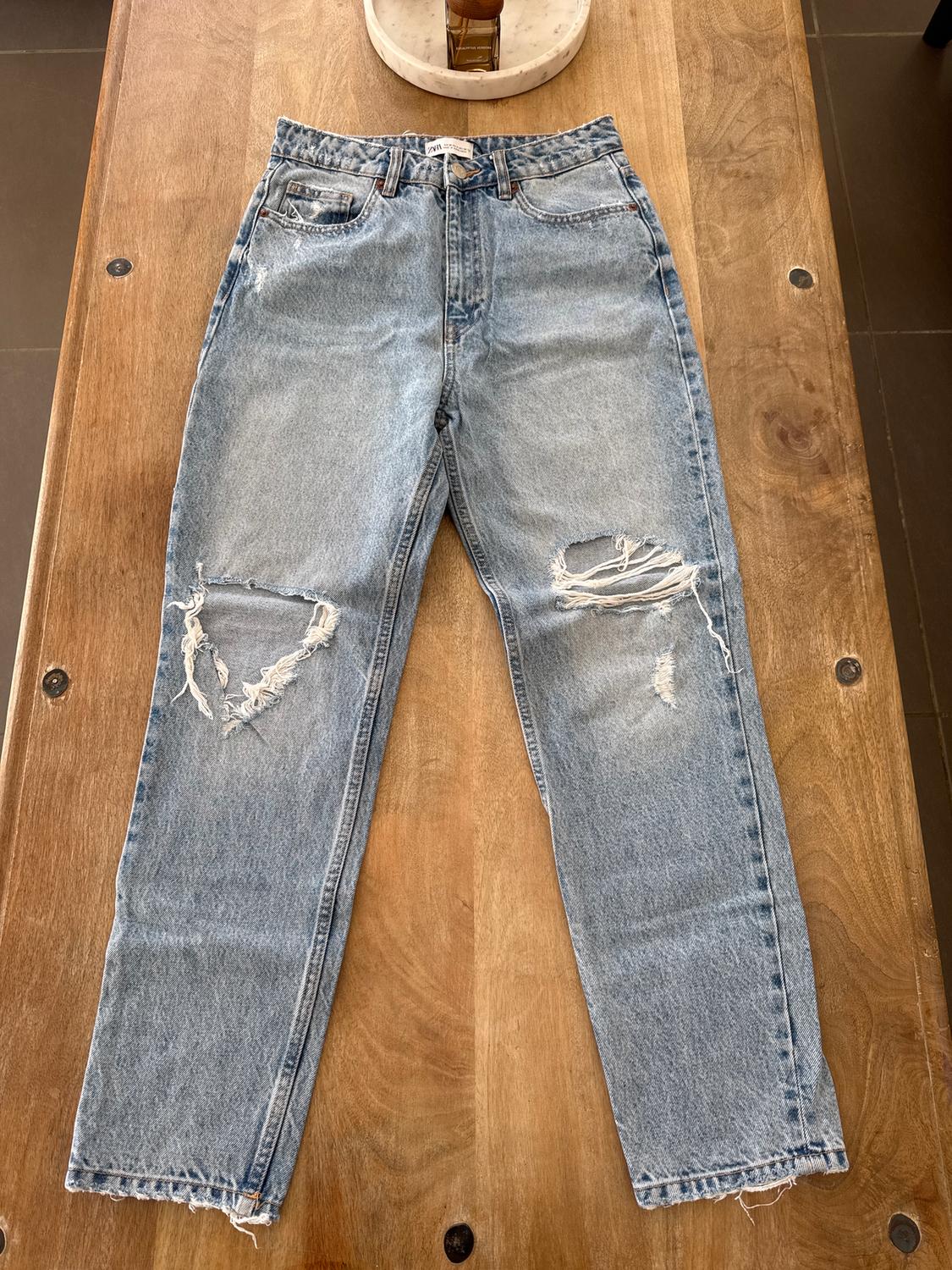 Distressed blue jeans with ripped knees, placed on a wooden table. Visible label on the waistband.