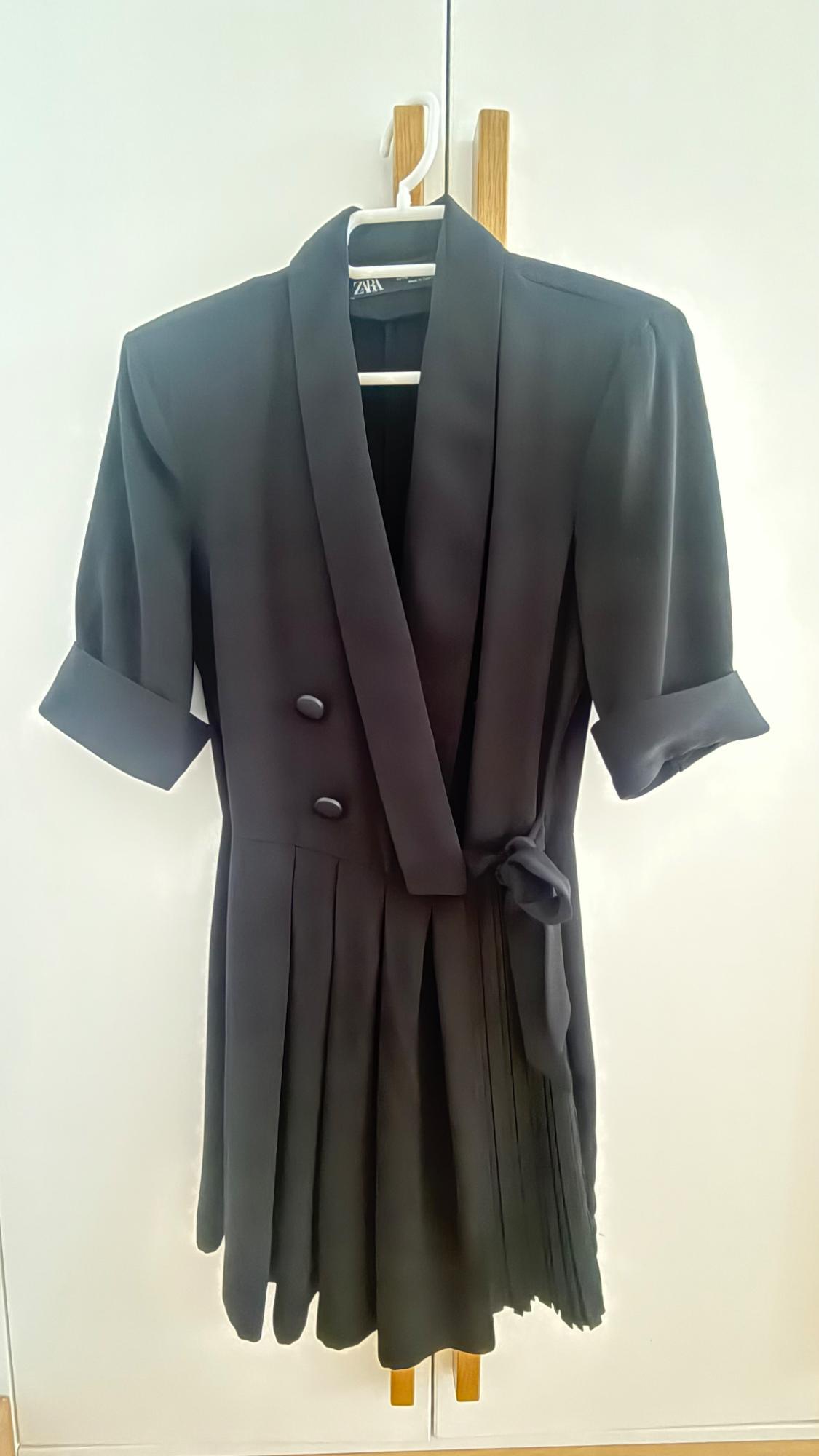 Black short-sleeved wrap dress with buttons and a pleated skirt, hanging in a white wardrobe with wooden handles. ZARA text.