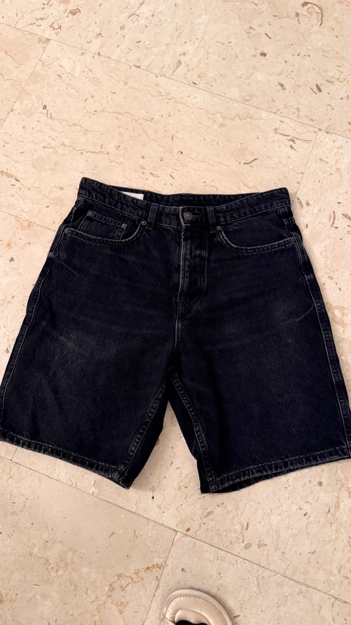 Black denim shorts on a light marble floor. Part of a white shoe visible in the bottom right corner.