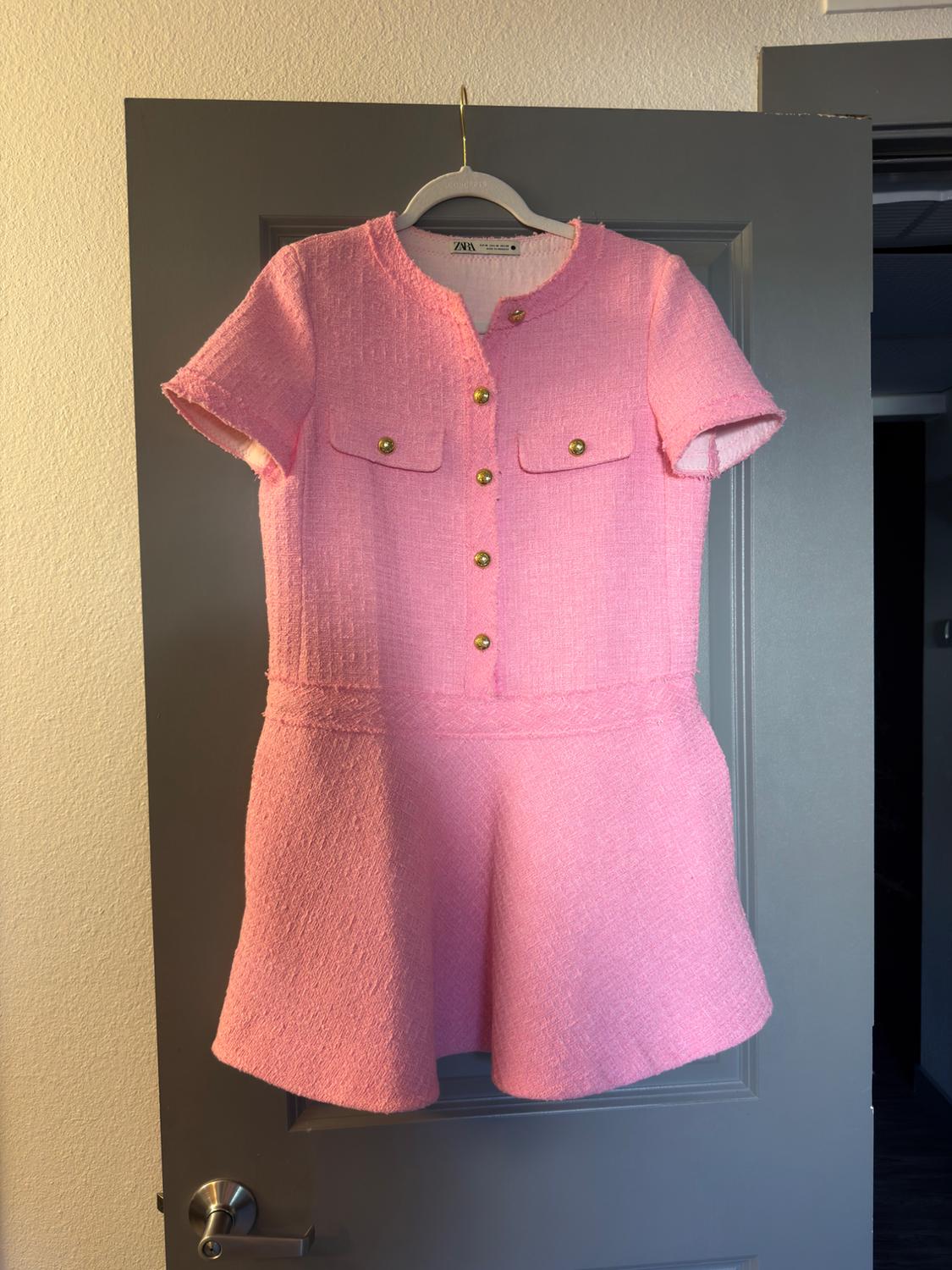 Textured pink short-sleeved dress with gold buttons and pockets, hanging on a grey door. Label with ZARA text.