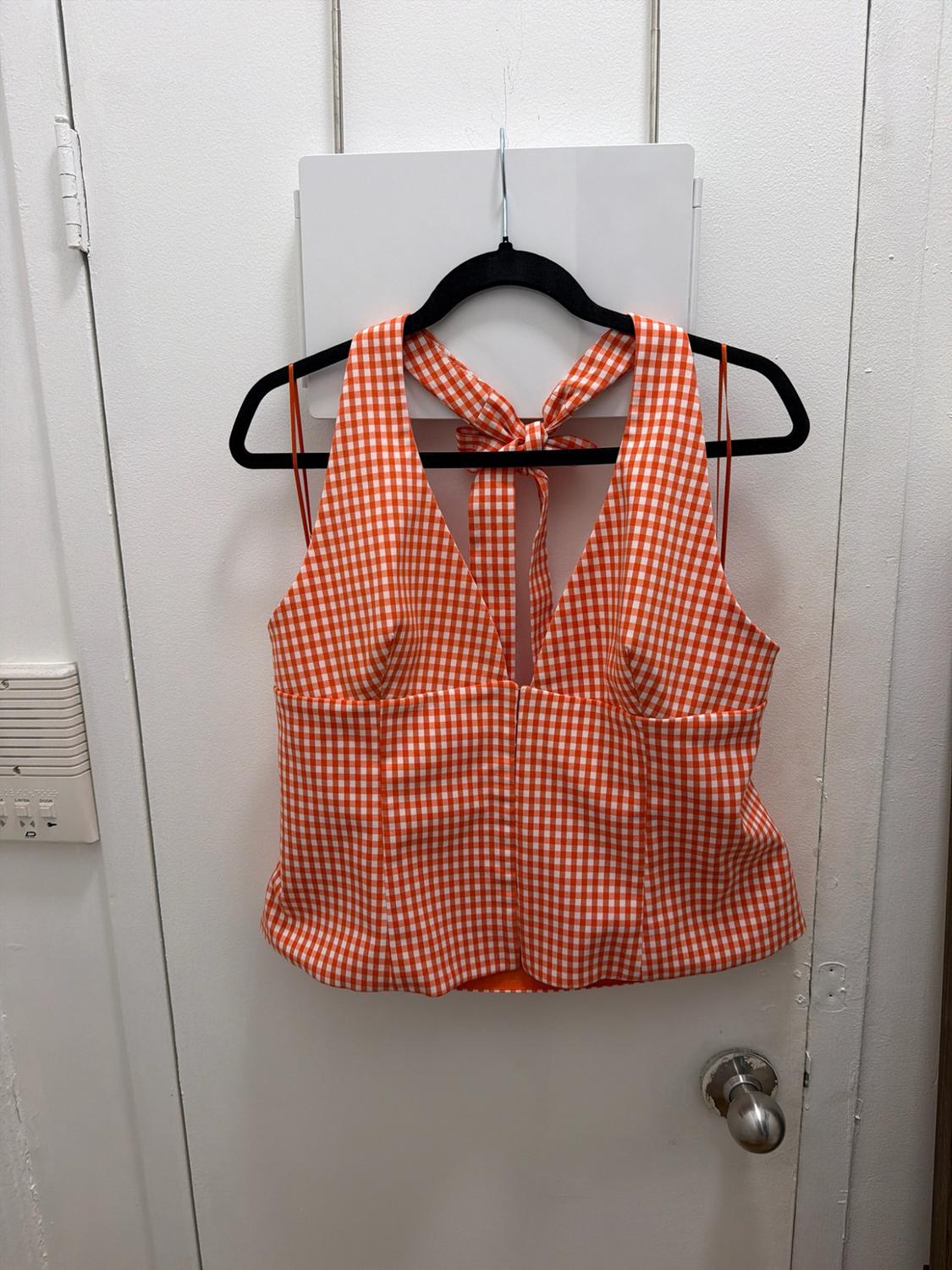 Top with an orange-and-white checkered pattern, a V-neckline, and a tie at the neck, hanging from a black hanger on a white door.