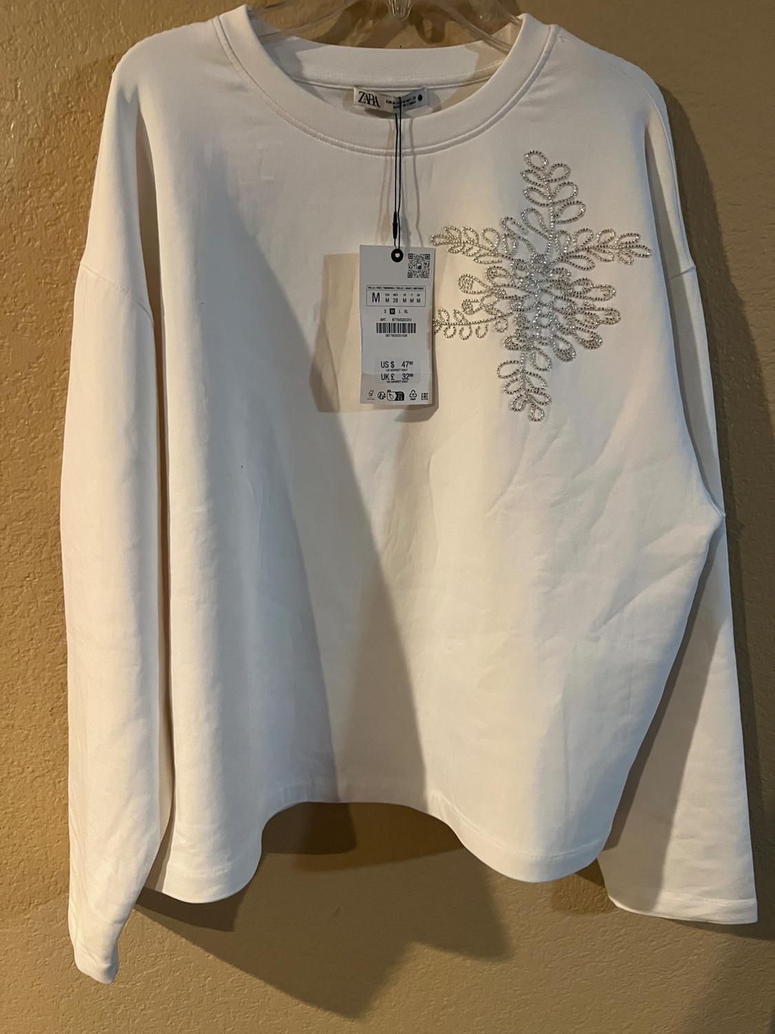 White sweatshirt with silver sequin snowflake design on the chest. Visible swing tag with sizing details.