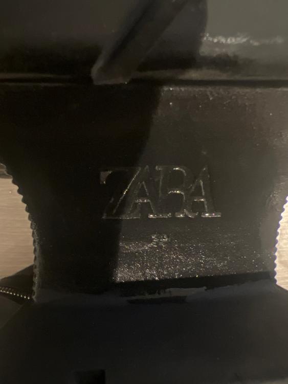 Close-up of the sole of a black shoe with the "ZARA" brand embossed. Dark background and rough texture.