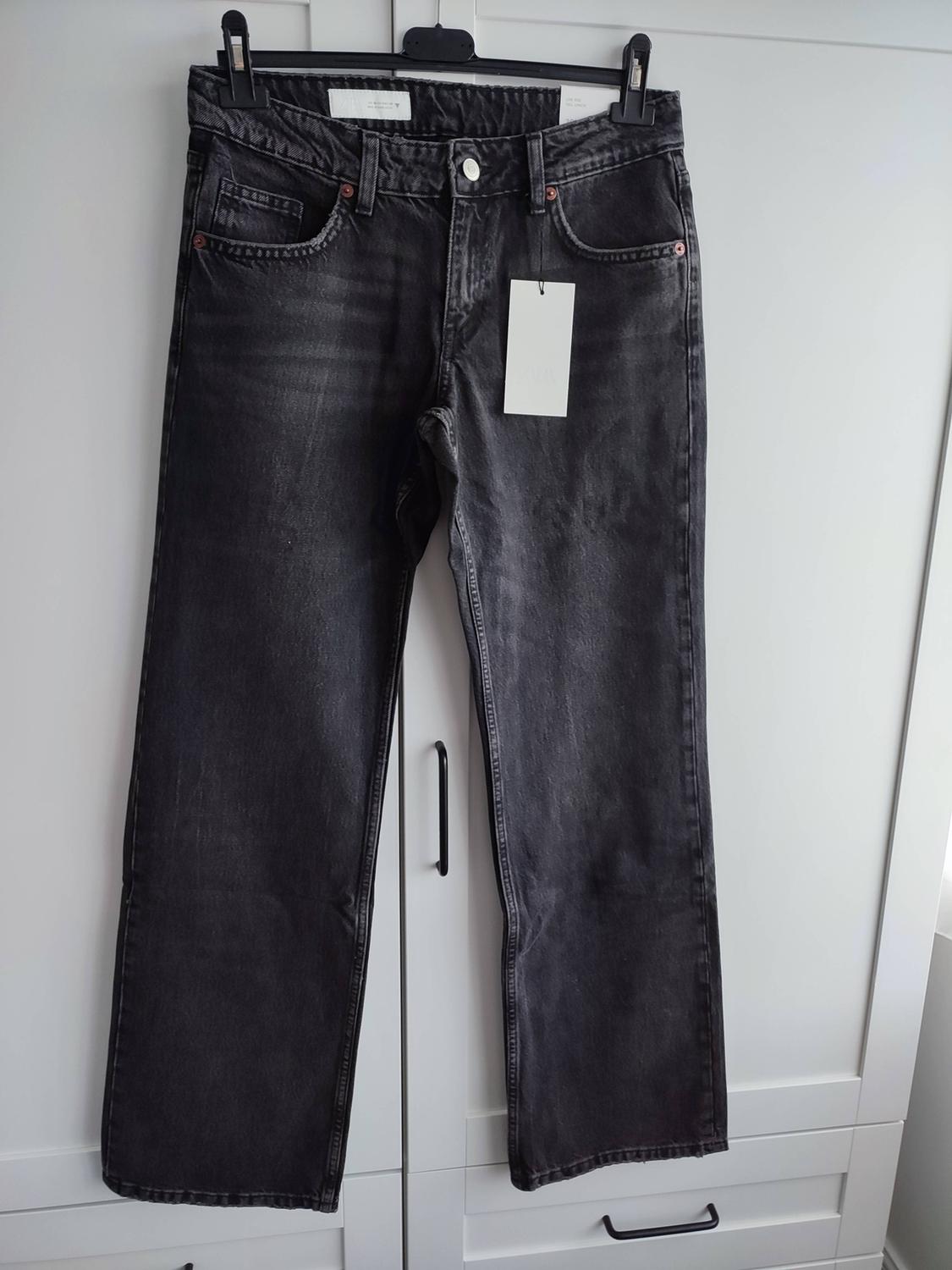 Distressed dark grey jeans hanging on a black hanger in front of a white wardrobe. It has a white label.