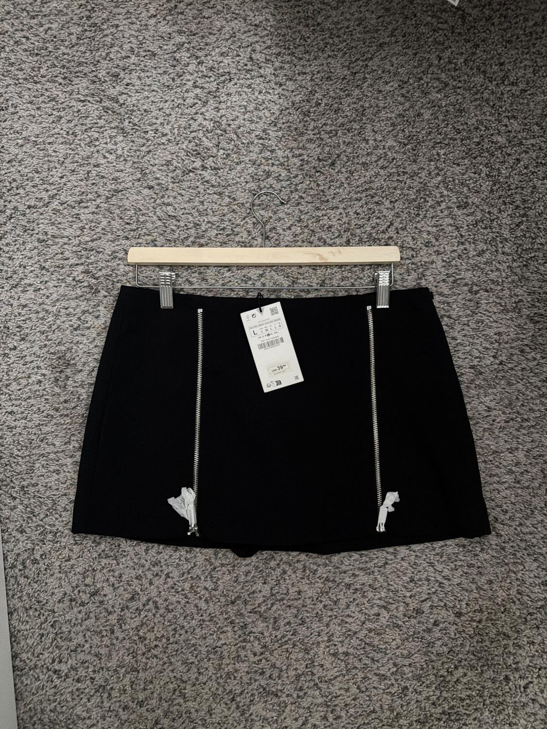 Black skirt with vertical zippers and hang tag, hanging on a wooden hanger over gray carpet.
