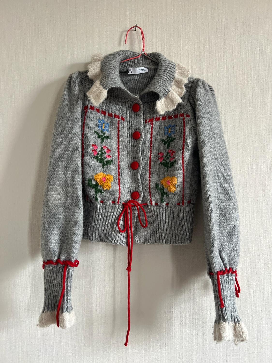 Gray knit cardigan from ZARA with a white ruffled collar and cuffs, floral embroidery, red buttons, and red bows at the waist, on a hanger.