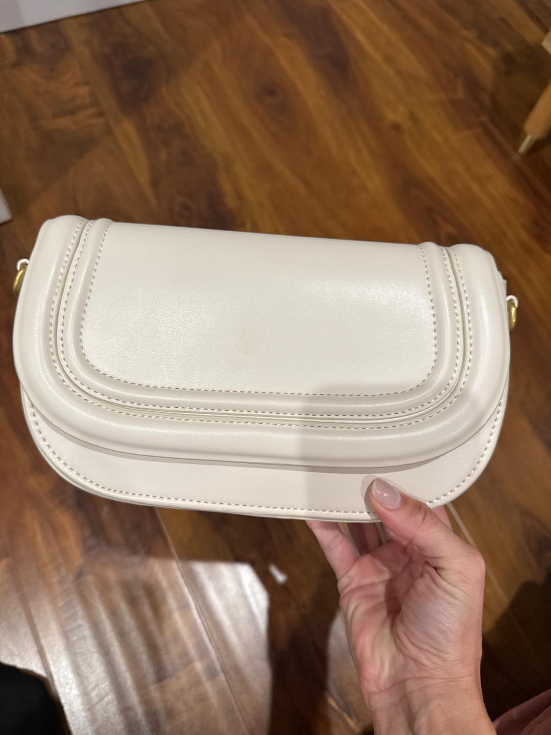 Hand holding a white handbag over a wooden floor. The handbag has a curved design with visible stitching.