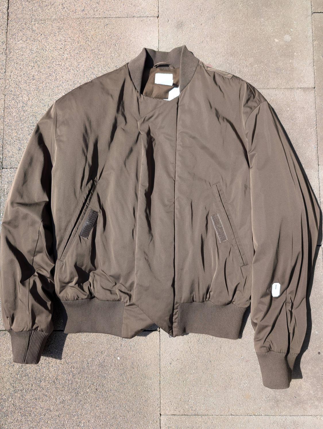 Brown bomber jacket on a tiled floor. It has elasticated cuffs and waist, with slanted pockets and a round neck.