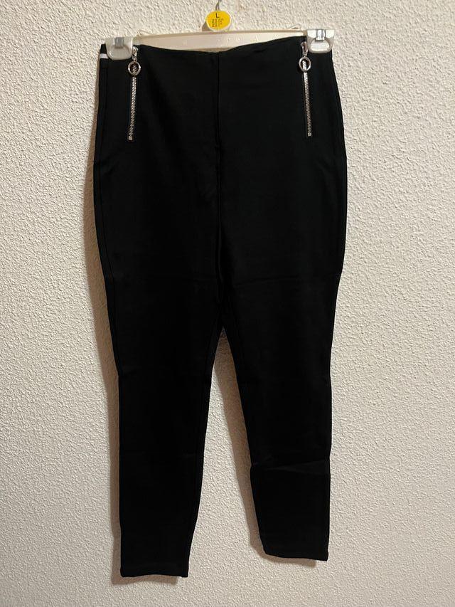 Black trousers hanging on a white hanger with two zips at the top, against a textured wall.