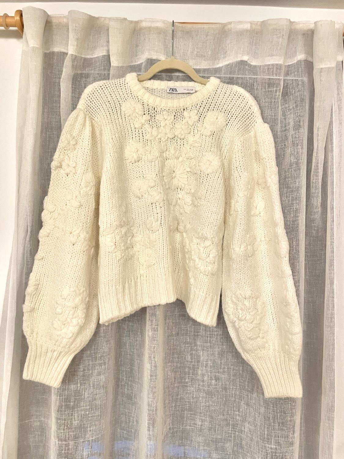 White chunky knit sweater with a raised floral design, hanging on a hanger in front of a sheer curtain.
