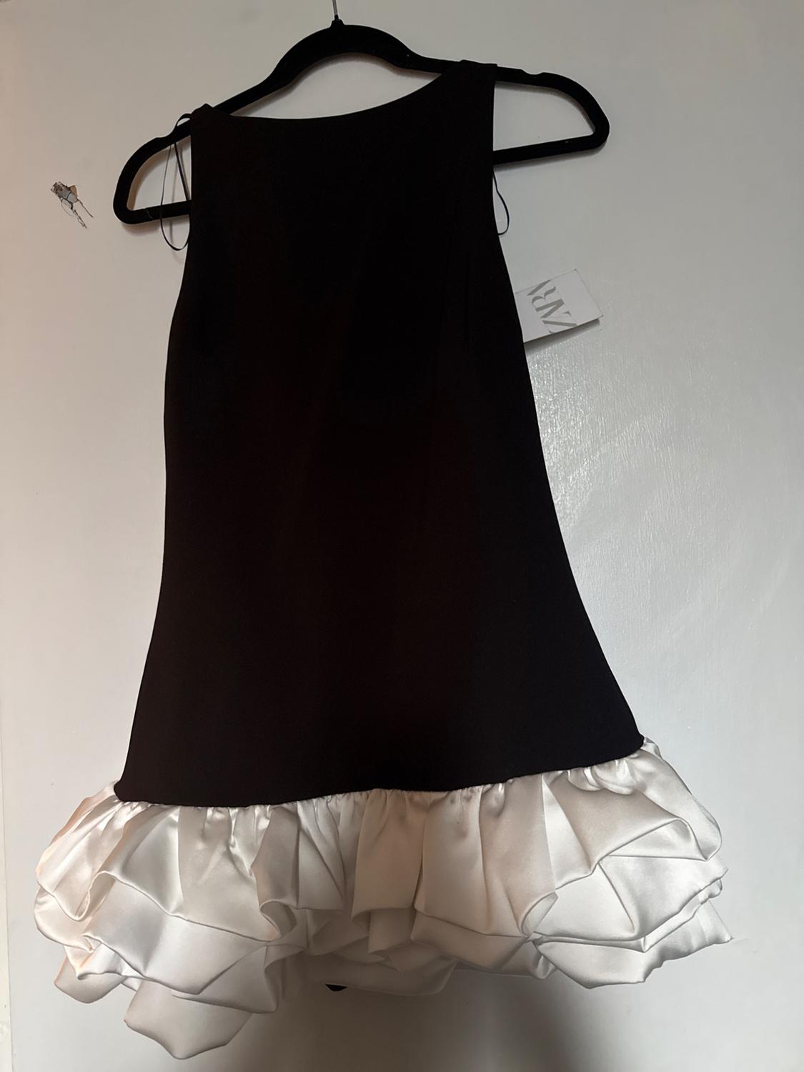 Sleeveless black dress with white ruffles on the hem, hanging on a black hanger against a white background. Tag visible.
