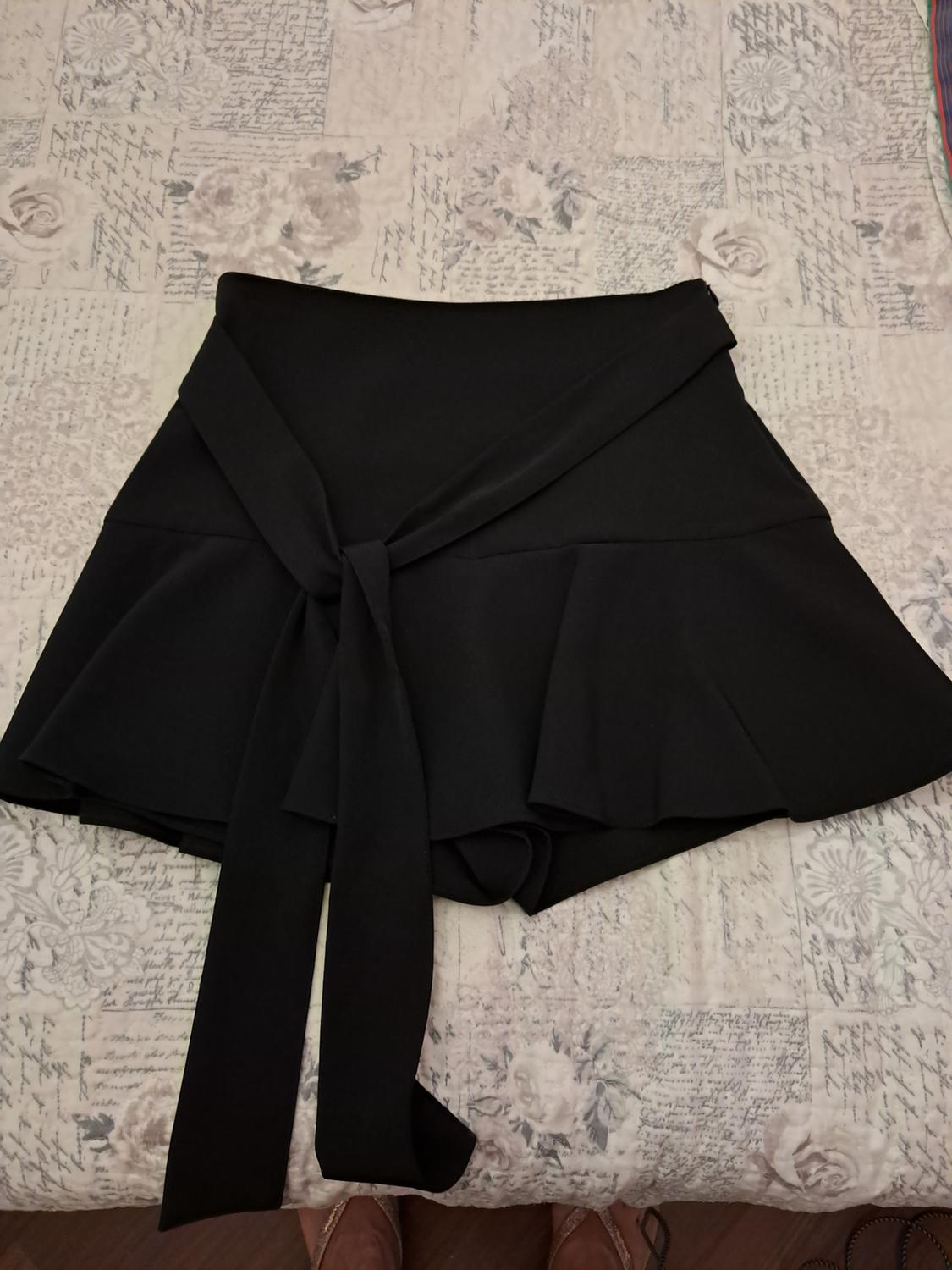 Short black skort with a flared cut and tied front bow, laid out on a bed with a light-coloured floral-patterned bedspread and text.