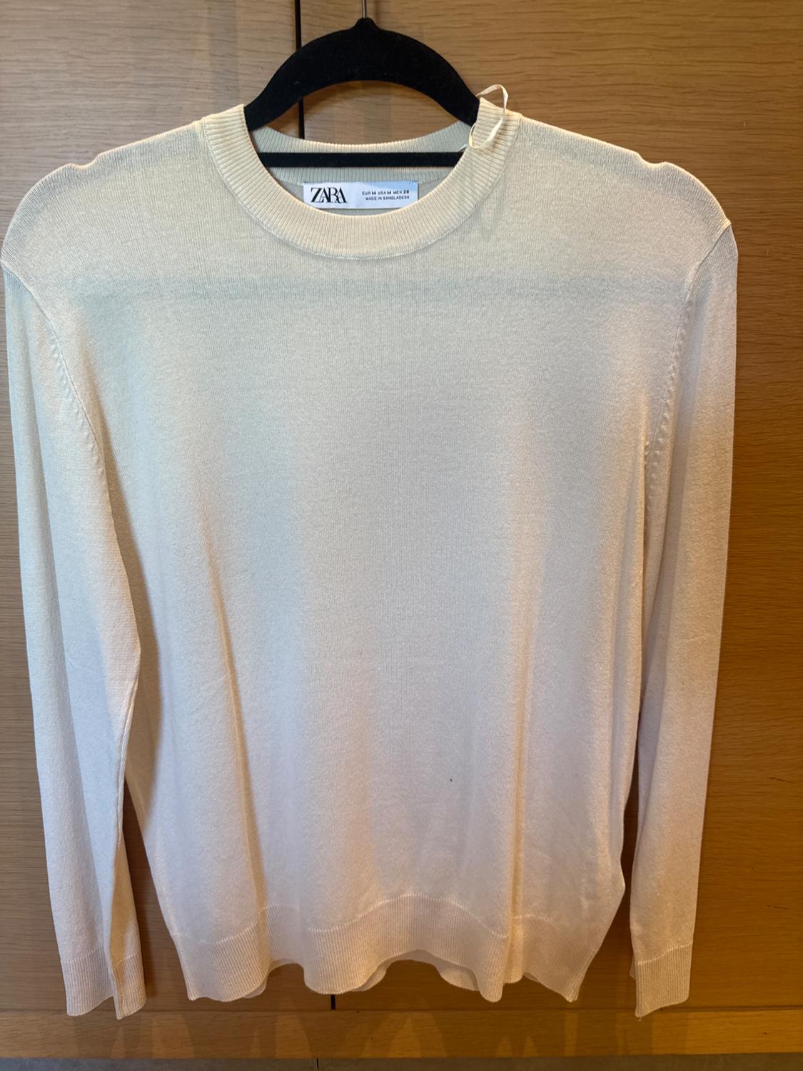 Beige long-sleeved jumper hanging on a black hanger, with a visible Zara tag, against a light wooden background.