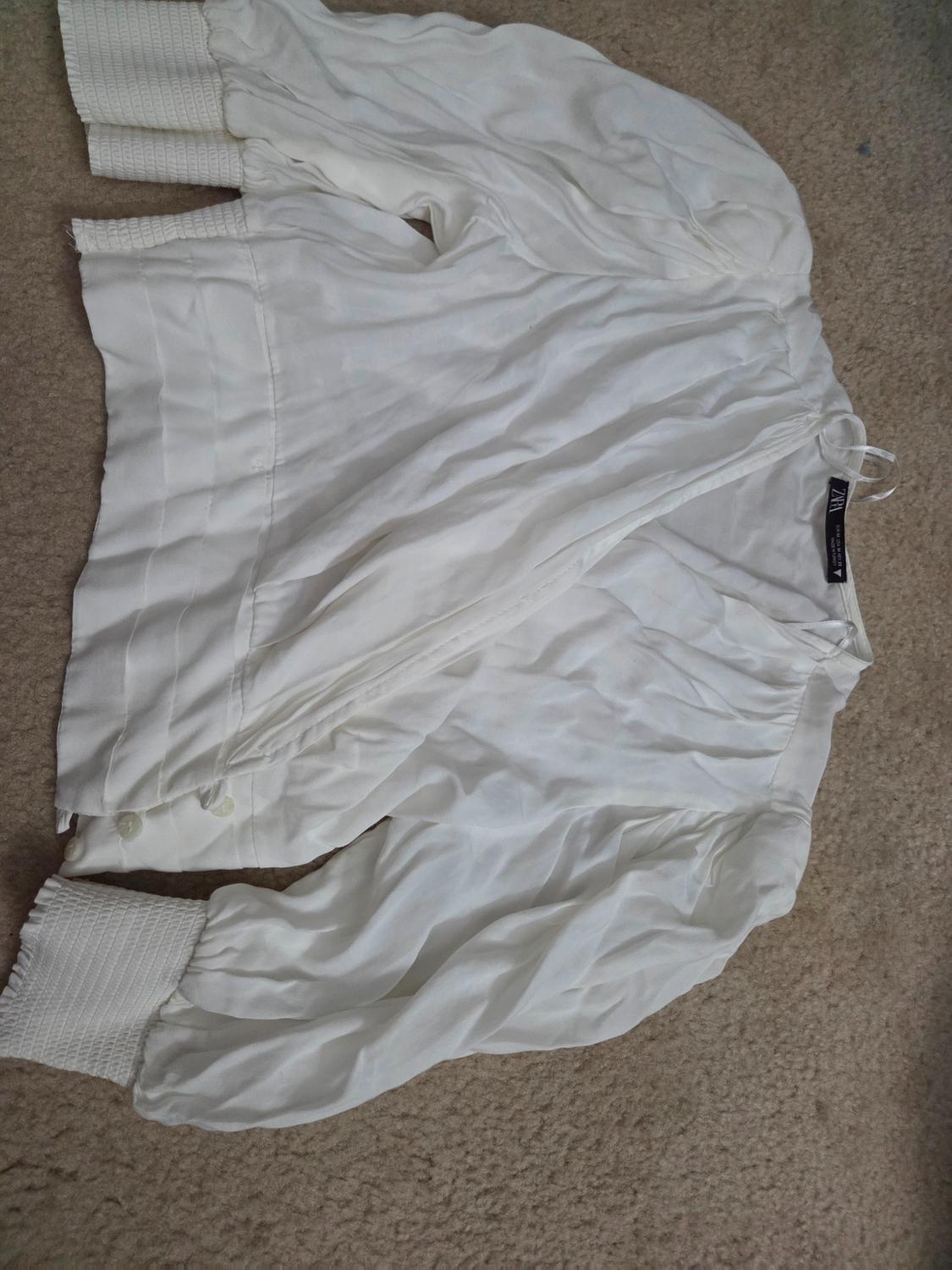 White long-sleeved jacket with elastic cuffs and buttons, placed on a beige rug. Visible tag on the collar.