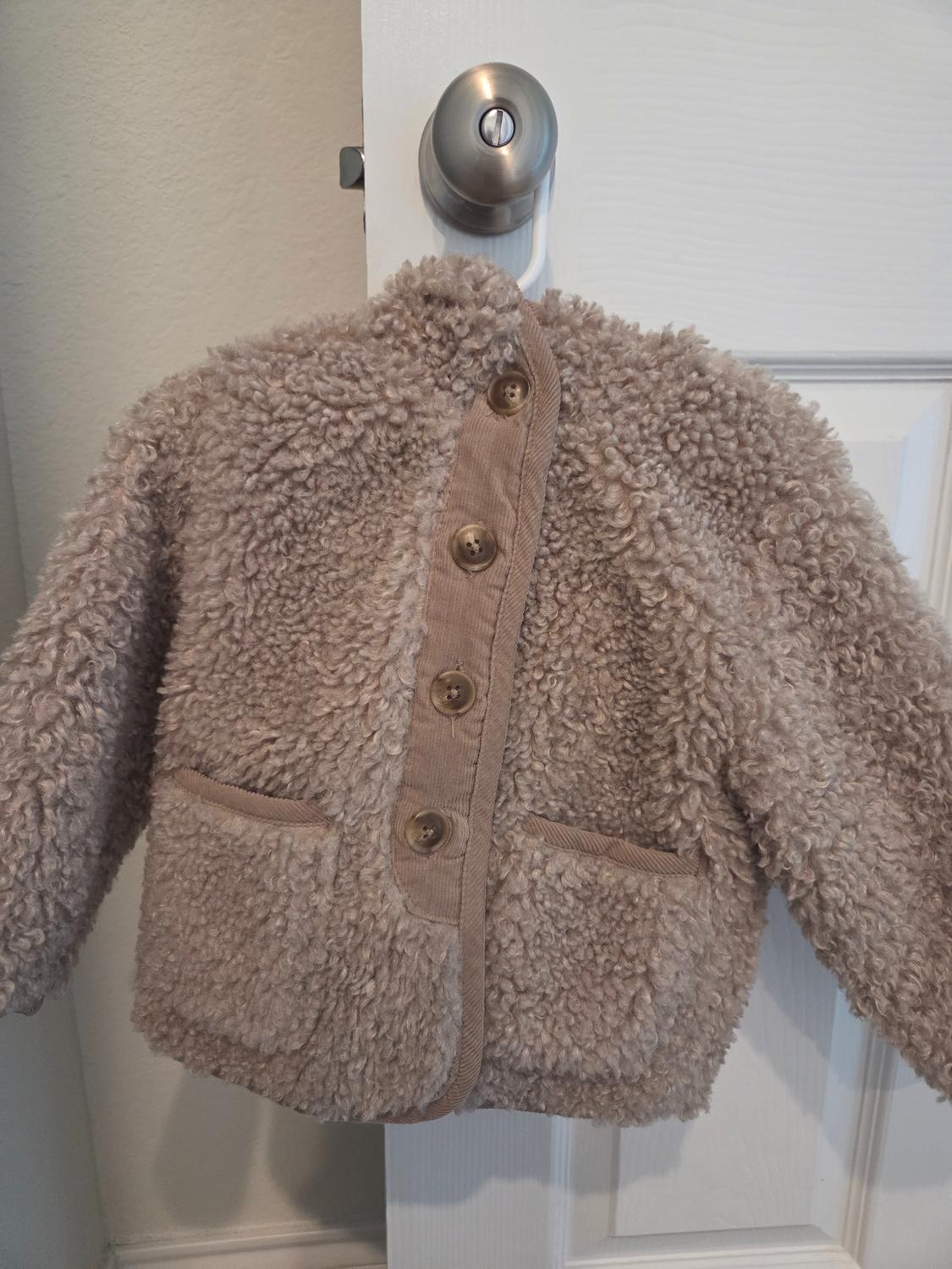 Beige wool jacket with brown buttons and two front pockets, hanging on a white door handle.