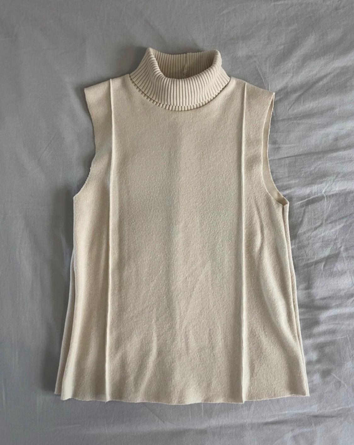 Cream sleeveless knitted top with a high neck and front vertical seams, laid on light grey fabric.