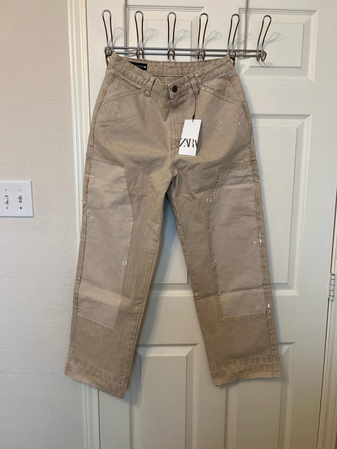 Beige utility-style trousers with white paint stains hanging on a door. Label with the text ZARA.