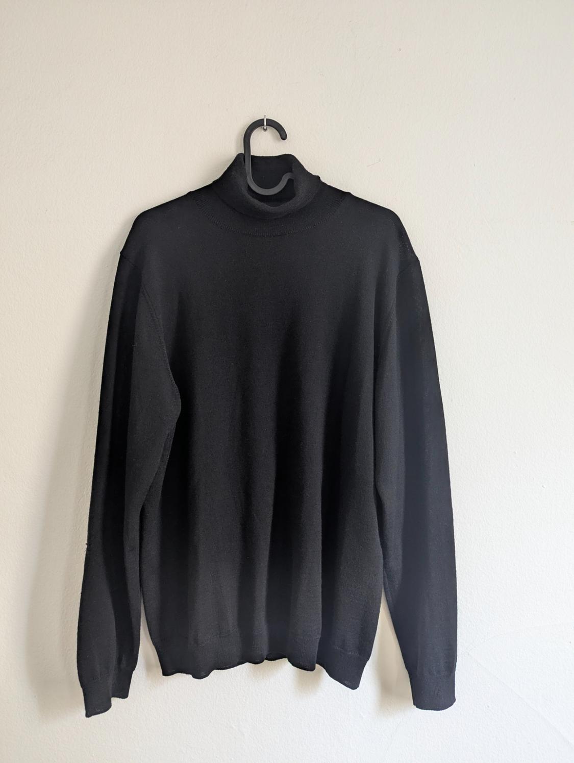 Black roll-neck jumper hanging on a hanger against a white wall.