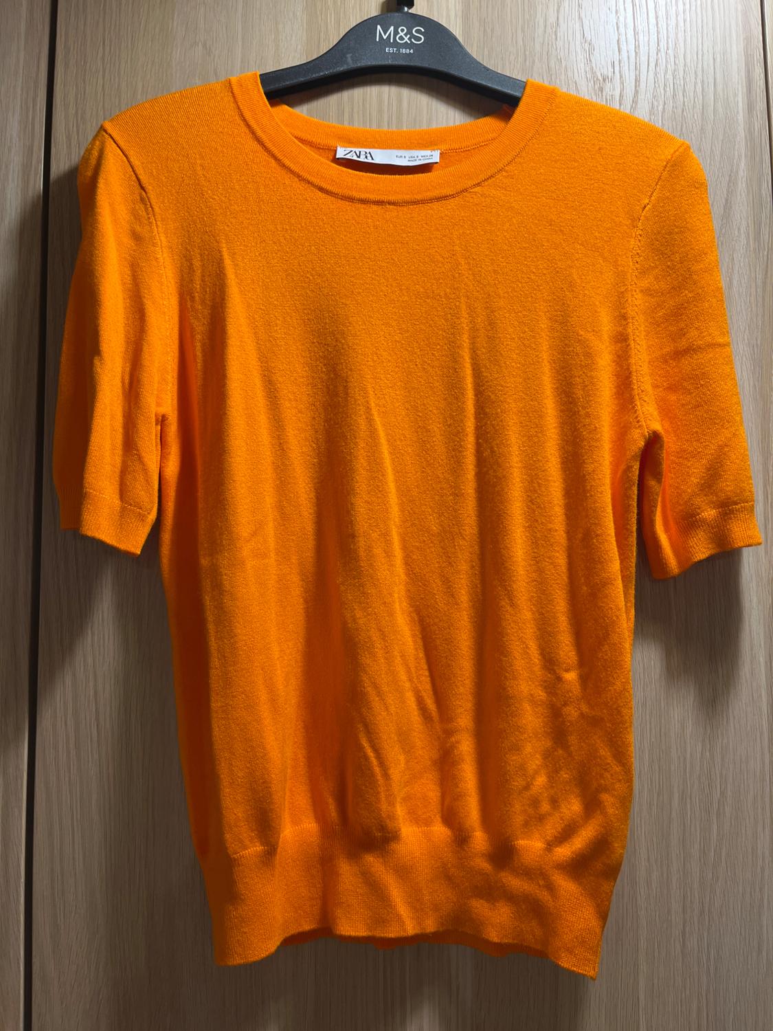 Short-sleeved orange jumper hanging on a black hanger with an "M&S" tag. Light wooden background.