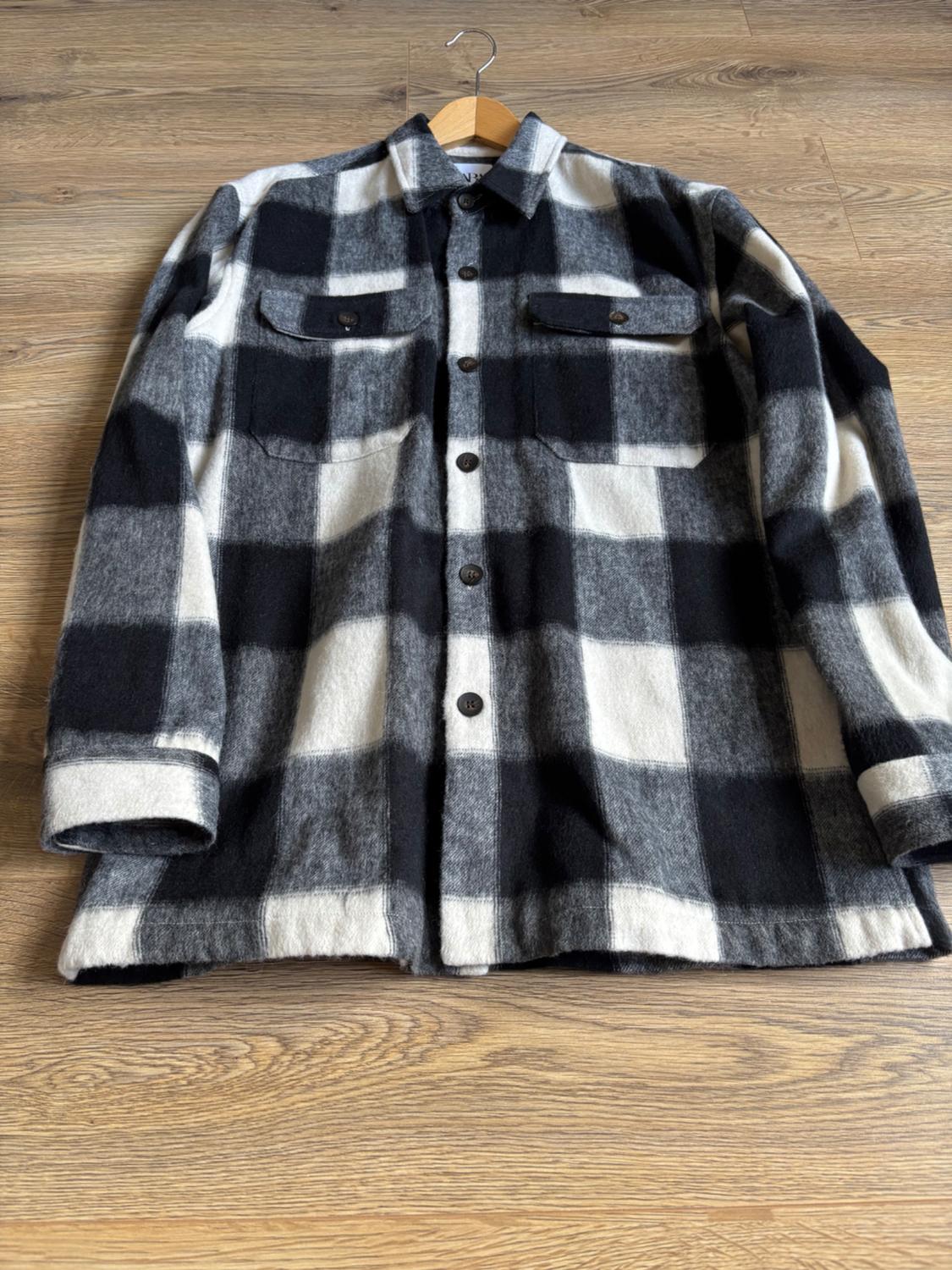 Black-and-white checked overshirt with buttons and front pockets, hanging on a coat hanger over a wooden floor.