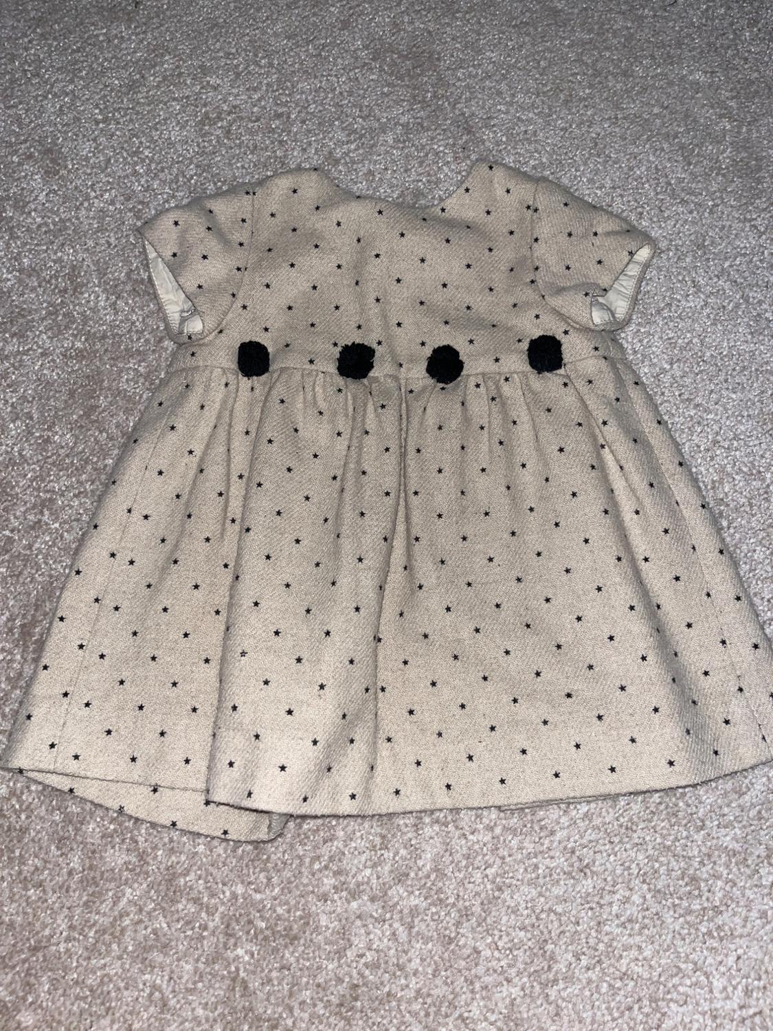 Child's beige dress with a small black star print and three black pom-poms at the waist, on a grey carpet.