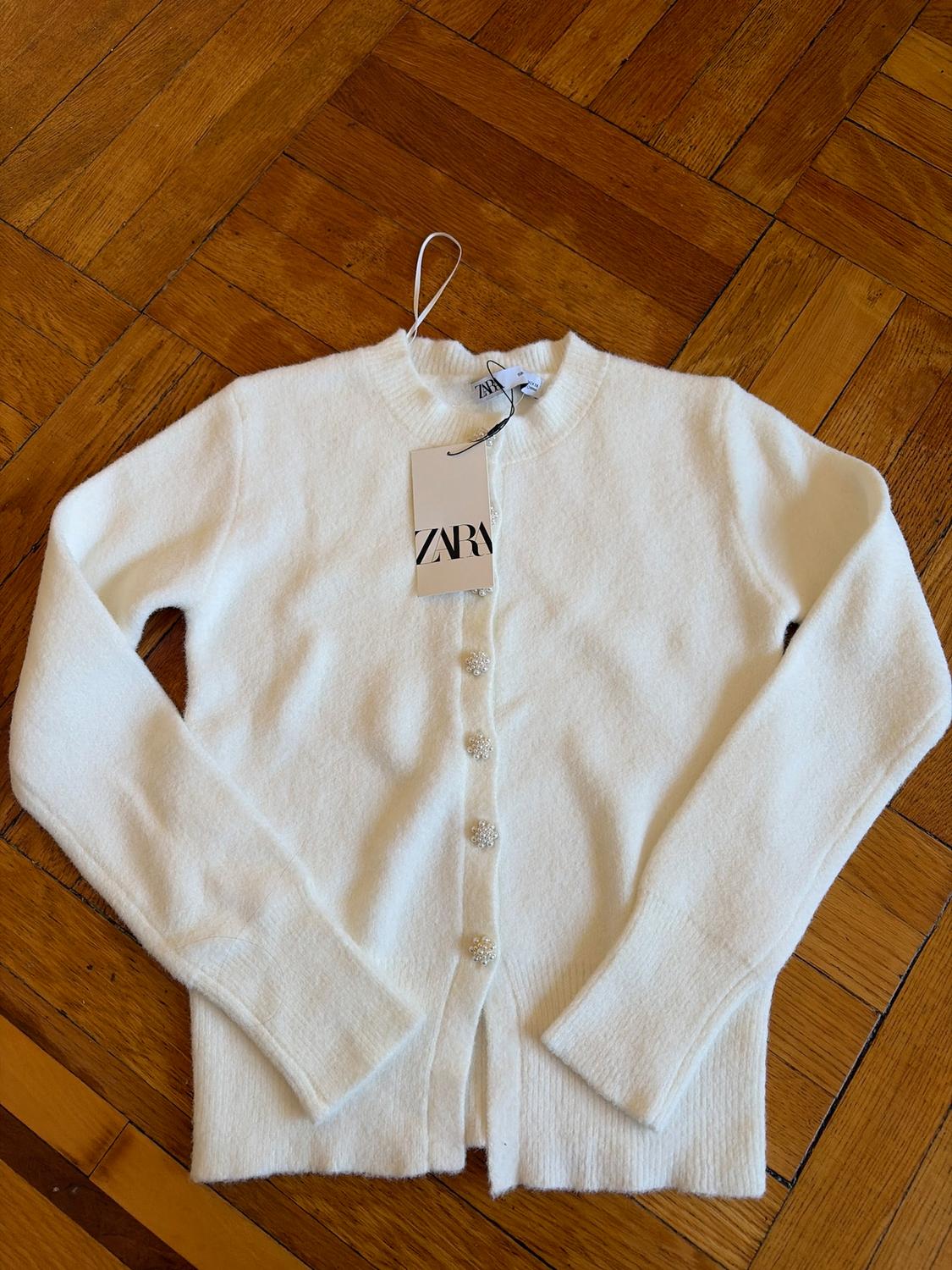 White long-sleeved cardigan with shiny buttons on a wooden floor. Visible label with the text "ZARA".