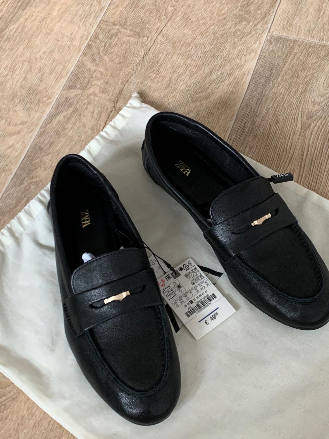 Black loafers with metallic detail on a white bag on a wooden floor. Text: ZARA, EAC, €49.95.