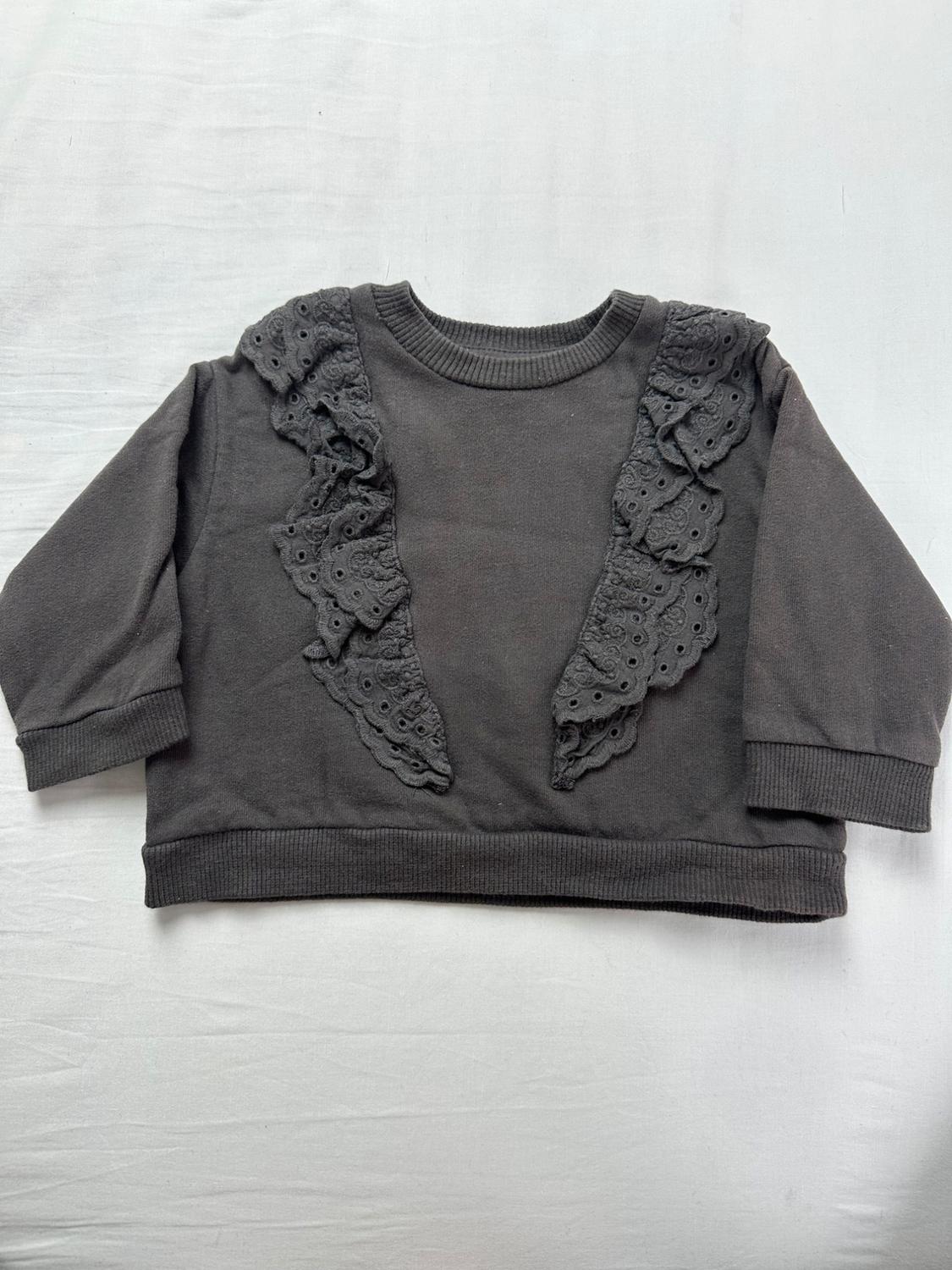 Dark gray kids' sweatshirt with long sleeves, ribbed cuffs, and decorative lace ruffles on the chest, on a white background.