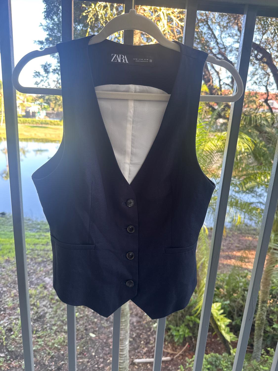 Black button-up vest hanging on a hanger with a visible tag, natural landscape background with trees and a lake.