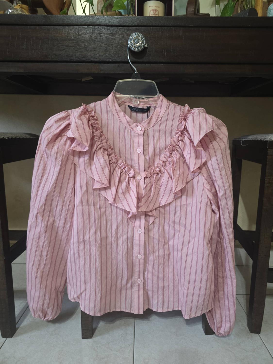 Pink striped blouse with ruffles, hanging on a hanger on a wooden piece of furniture. Text: ZARA, L.