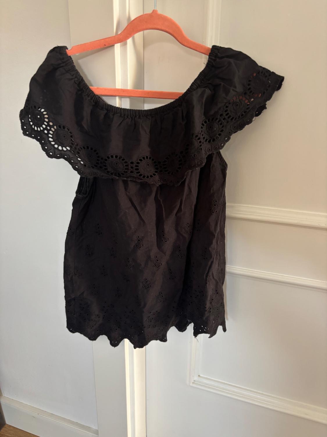 Black blouse with a bateau neckline and lace detailing hanging on an orange hanger over a white door.