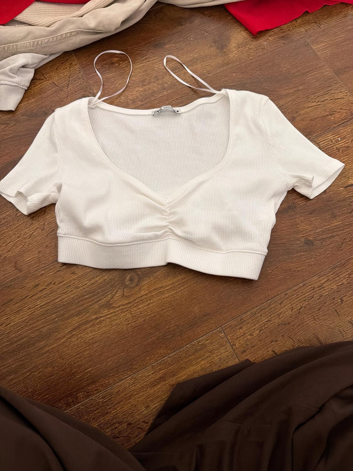 White short-sleeved V-neck top on a wooden floor. Around it, garments in beige and red tones. Visible label.