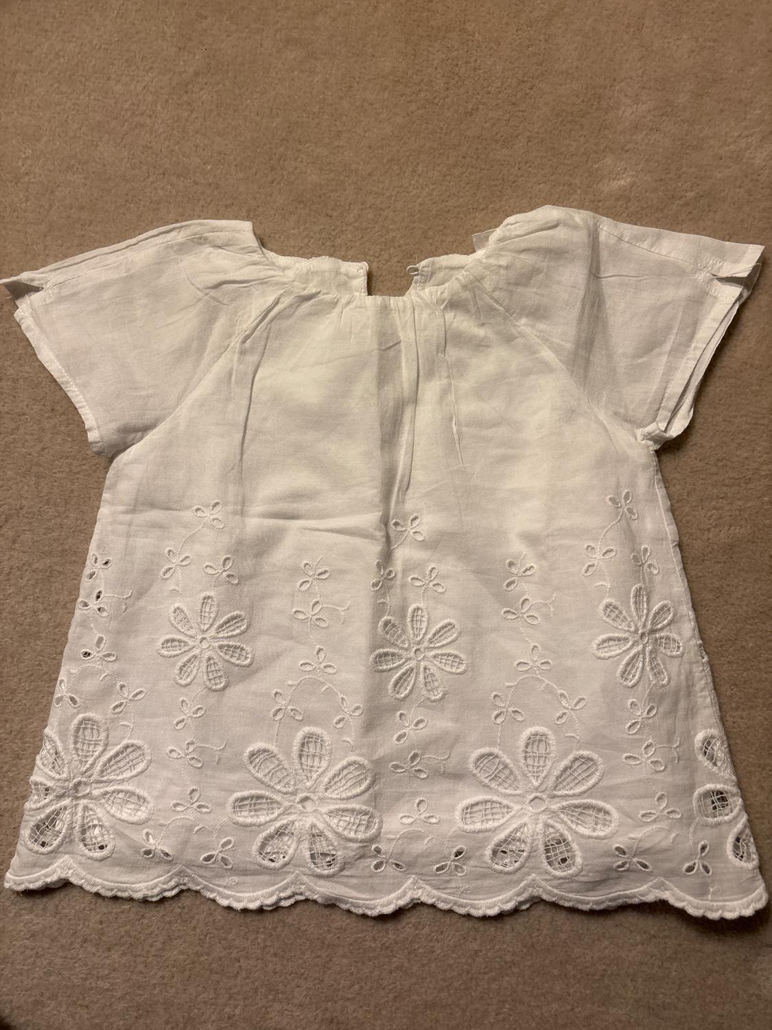 White short-sleeved blouse with floral embroidery and openwork details at the bottom, on a beige background.