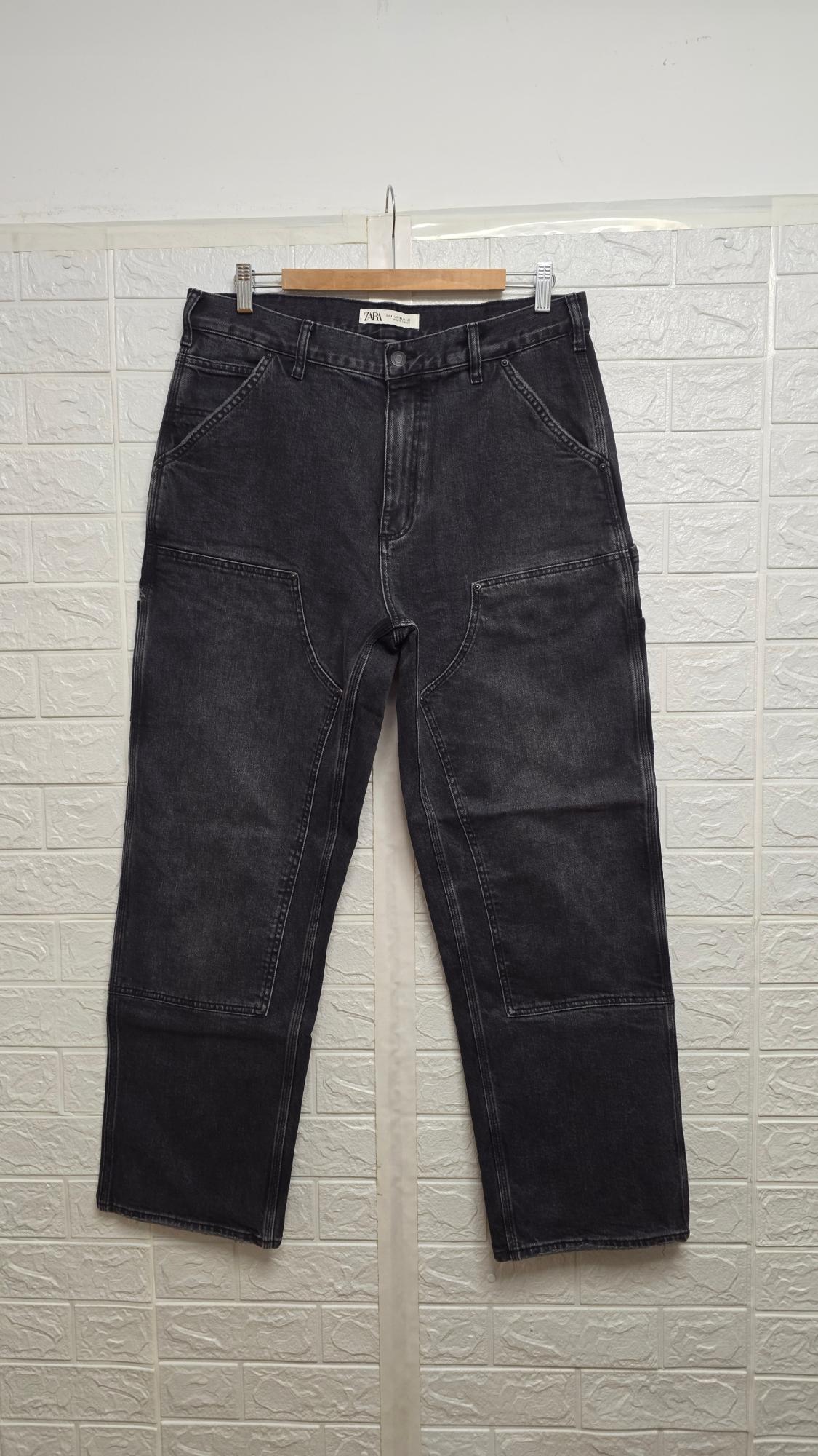 Black jeans hanging on a wooden coat hanger, with visible seams, against a white brick textured wall background.