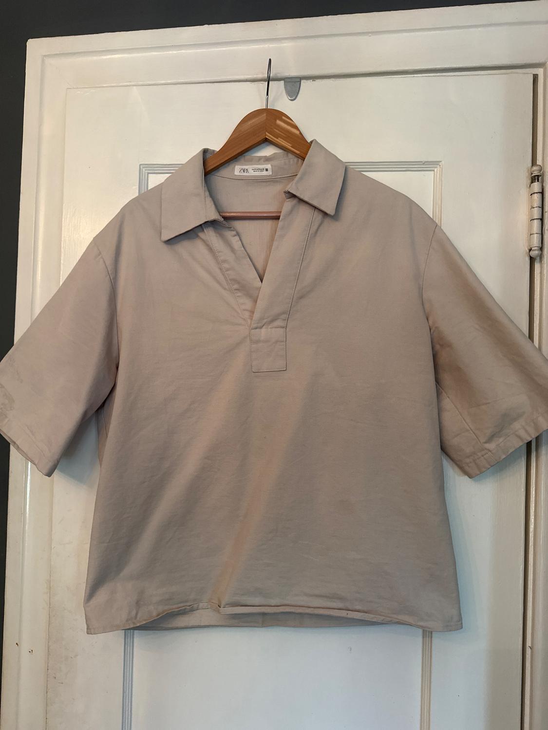 Beige short-sleeved shirt hanging on a wooden hanger over a white door. Visible label with text "ZNR".
